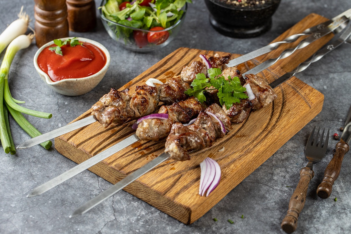 Das Bild zeigt sechs Schaschlikspieße, von denen drei auf einem Holzbrett liegen und mit roten Zwiebeln und Petersilie dekoriert sind. Die anderen drei Schaschlikspieße sind ebenfalls fertig gegrillt und mit roten Zwiebeln und Petersilie dekoriert. Daneben liegt eine kleine Schüssel mit Ketchup und eine kleine Schüssel mit gemischtem Salat..