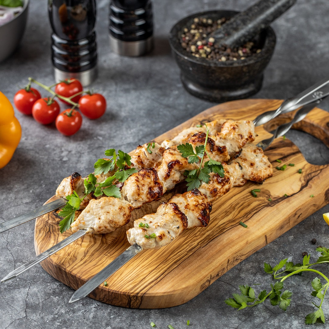 "Fertiges Hähnchen-Schaschlik original auf edelstahlspießen präsentiert auf einem Holzbrett mit dekorativen Tomaten und frischer Petersilie.