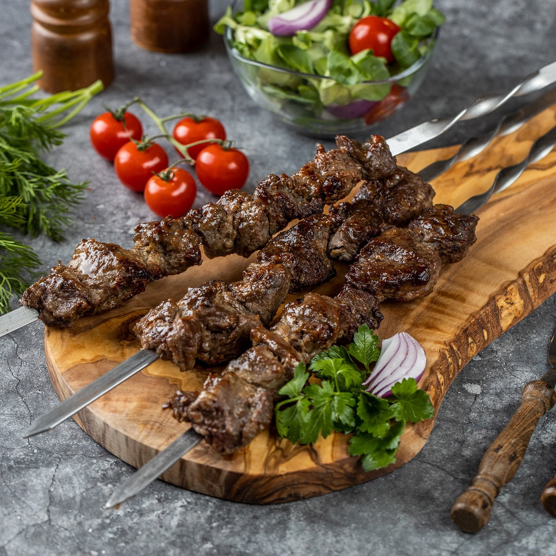 "Fertiges Lamm-Schaschlik auf edelstahlspießen präsentiert auf einem Holzbrett mit Tomaten, Petersilie und roten Zwiebeln.