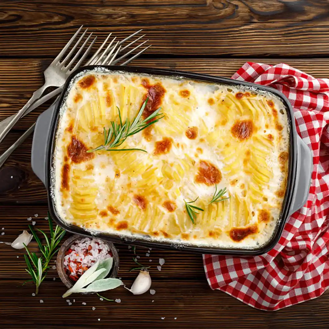 Kartoffelgratin in Auflaufform, garniert mit Kräutern und angerichtet auf einem Holztisch mit Besteck, Salz, Knoblauch und einem Küchen Handtuch. Serviervorschlag.