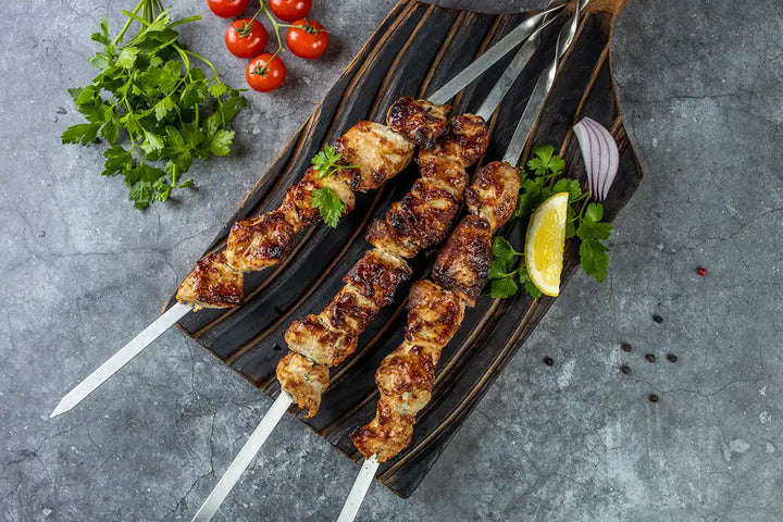 Pute Schaschlik Original auf einem Holzbrett angerichtet, garniert mit Zitrone, Petersilie und Zwiebel. Dekoriert auf einer Arbeitsplatte mit Cherrytomaten, Salat, Paprika und Gewürzmühlen aus Holz. Serviervorschlag.