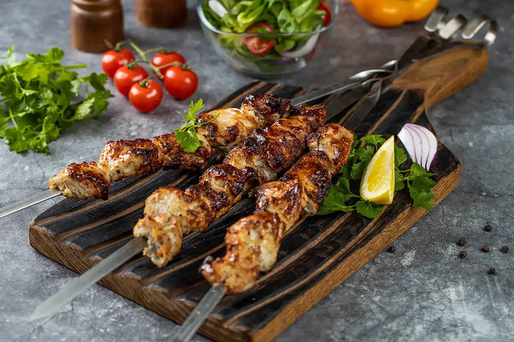 Pute Schaschlik Original auf einem Holzbrett angerichtet, garniert mit Zitrone, Petersilie und Zwiebel. Dekoriert auf einer Arbeitsplatte mit Cherrytomaten, Salat, Paprika und Gewürzmühlen aus Holz. Serviervorschlag.