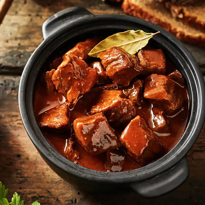 Rindersaftgulasch in Sauce serviert im Eintopf mit Lorbeerblatt auf einem Holzbrett, mit Brot im Hintergrund. Serviervorschlag.