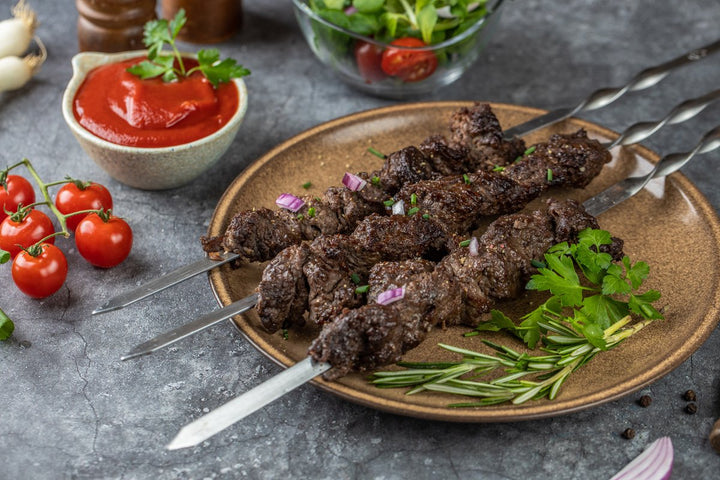 fertig gegrilltes Rinder-Schaschlik Original aufgespießt auf einem Teller liegend und mit roten Zwiebel, Petersilie und Rosmarien dekoriert