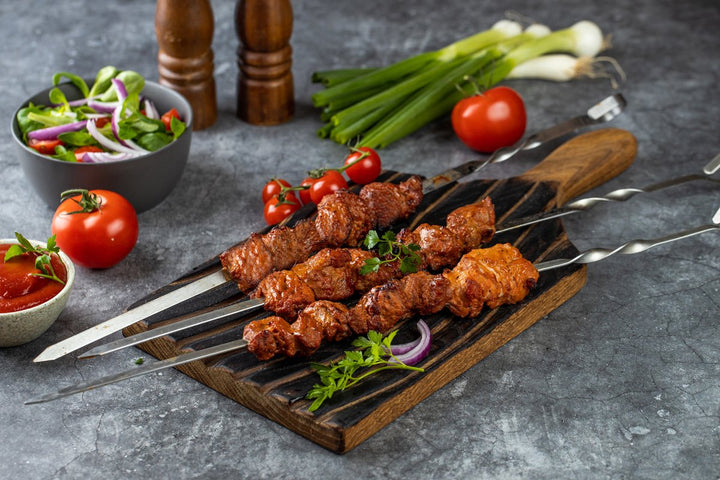 fertig gegrilltes Schweine-Schaschlik Tomate auf einem Holzbrett dekoriert mit Petersilie und roten Zwiebel. Links im Hintergrund steht eine kleine Schüssel Salat